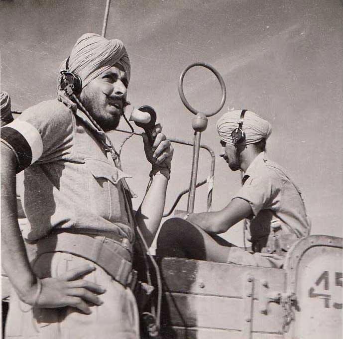 Two soldiers in turbans in an open vehicle; one speaks into a radio handset, a wireless aerial frame behind him.