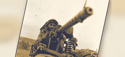 A gunner in desert headdress behind a heavy machine gun mounted on a truck, the barrel raised against an empty sky.