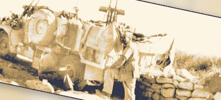 An LRDG truck under camouflage netting beside a dry-stone wall, twin Vickers K guns mounted fore and aft; one man sits by the rear wheel, another stands beside him.