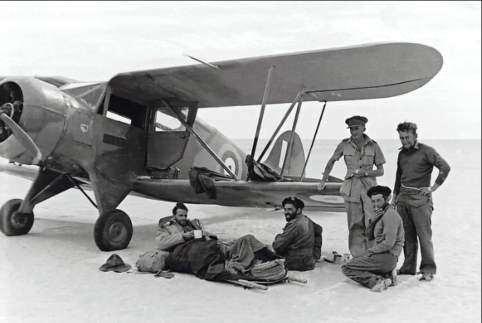 A Waco cabin biplane on a desert landing ground, five men beside it, one lying on the sand waiting to be flown out