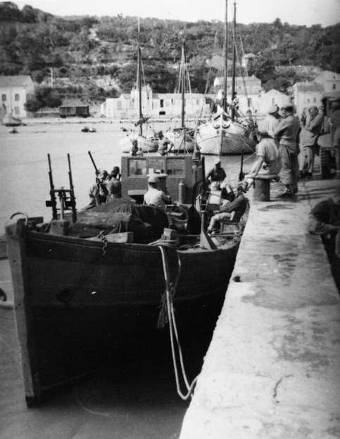 A small armed craft lying bow-on at a quay with men aboard, masted vessels and white houses under a wooded hill behind