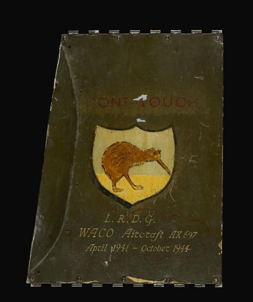 An olive-green aircraft panel painted with a kiwi on a shield, the words DONT TOUCH above it and an inscription below