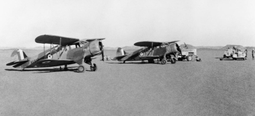 Two Waco cabin biplanes parked on a flat desert landing ground, with trucks and men beyond them